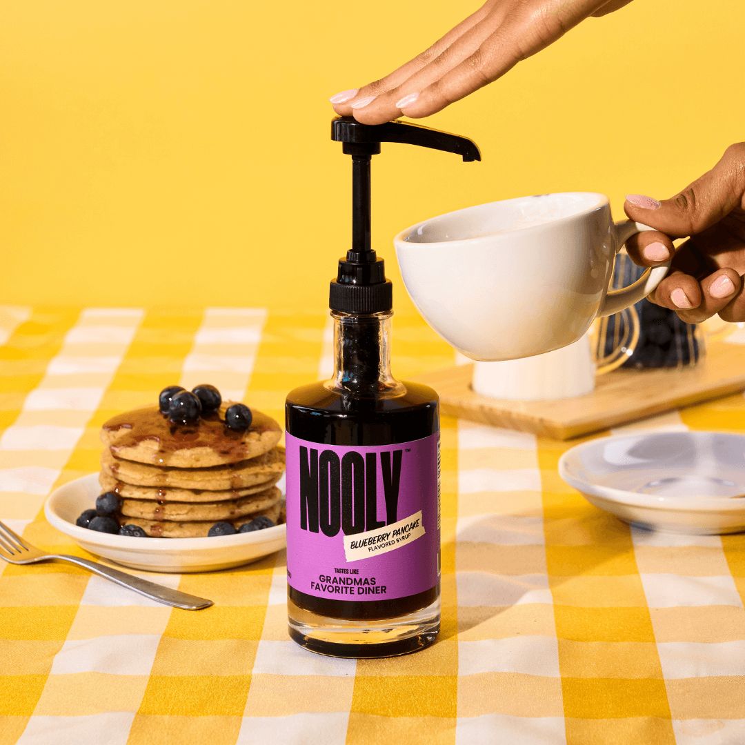 Bottle of Nooly with a syrup dispensing pump attached to the top and a hand pressing down a pump of syrup into a coffee cup